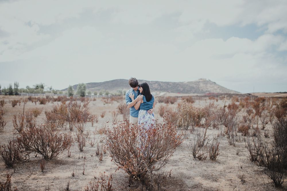 A romantic couple embraces in a desert setting with mountains and brush in the background.