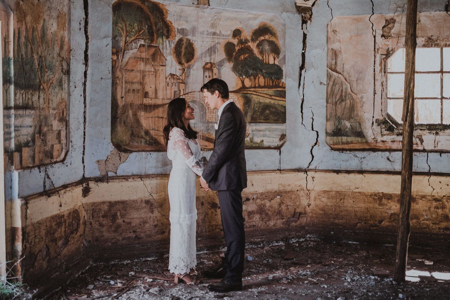 Couple in wedding attire standing in historic room with painted wall murals.