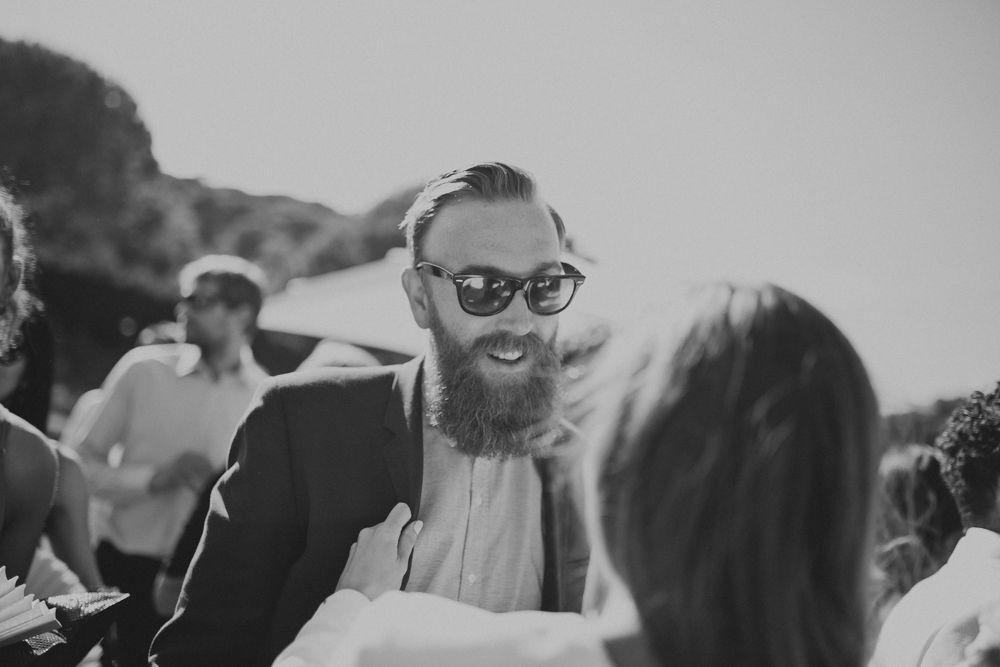 Wedding guest with beard and glasses smiles during outdoor celebration.