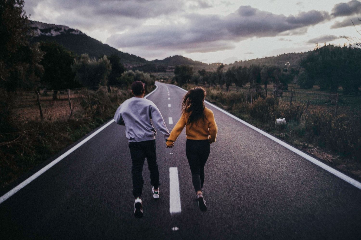 A couple holding hands while walking down the center of an empty mountain road.
