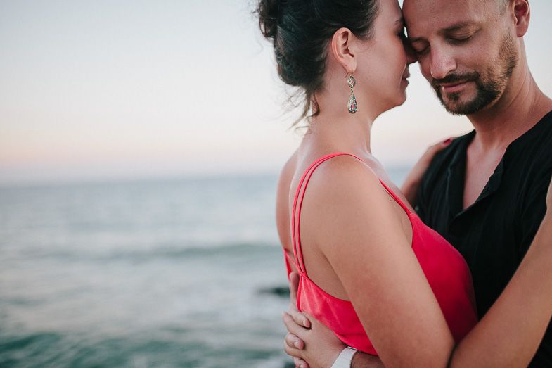 A romantic couple embraces near the ocean at sunset, with the woman wearing a red dress.