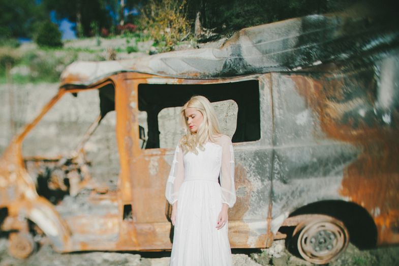 Woman in white dress standing in front of burnt out vintage vehicle.