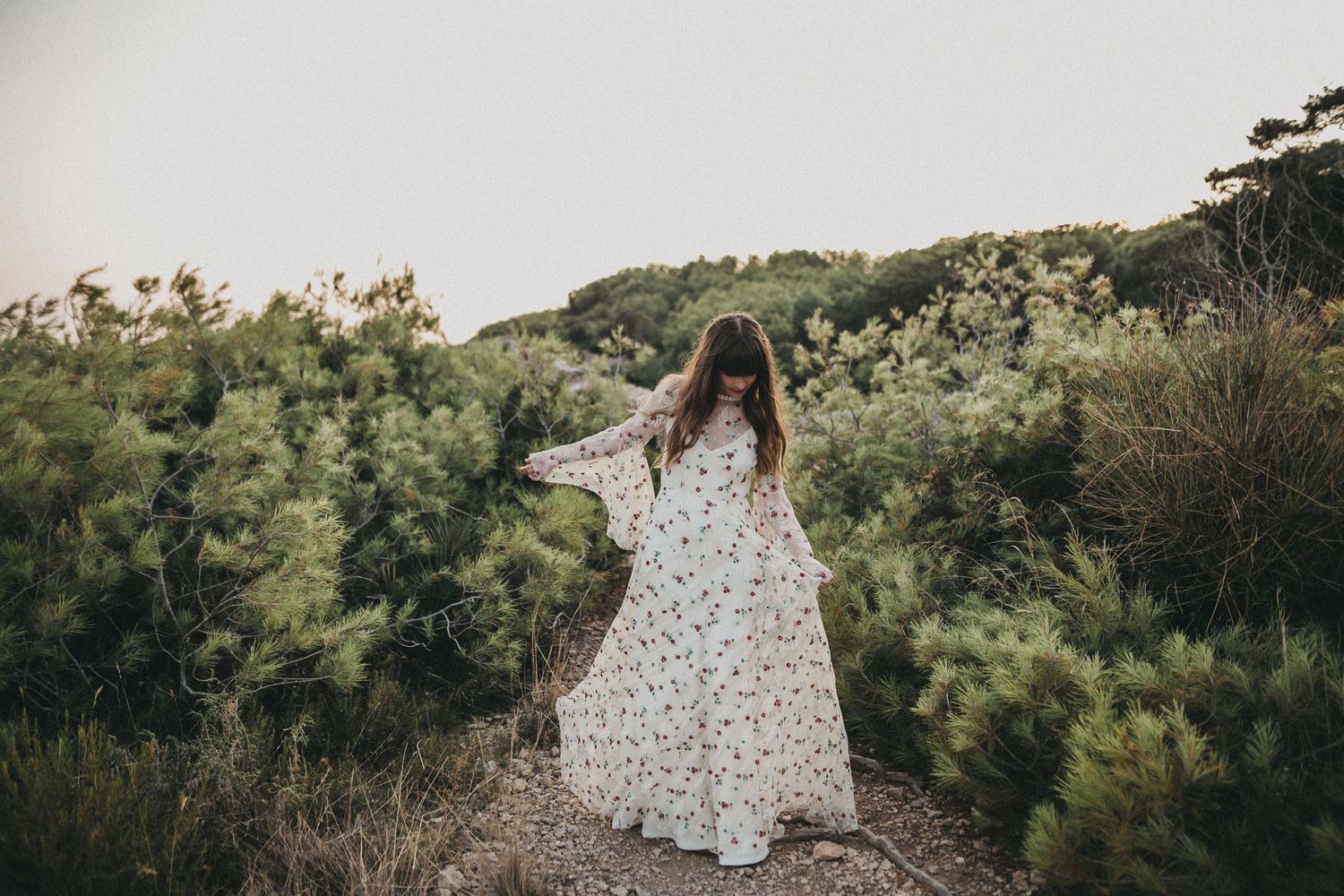 A flowing white floral dress twirls against a hillside backdrop with pine trees at sunset.