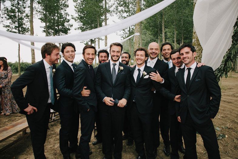 Groomsmen in dark suits pose together at an outdoor forest wedding venue.