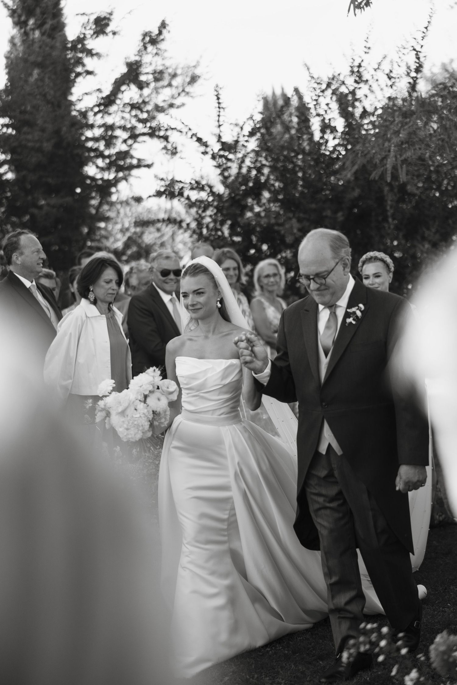 Black and white photo of a bride in a strapless gown walking down the aisle with her father at an outdoor wedding.