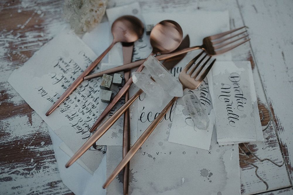 Copper cutlery and place cards arranged on rustic white wooden surface with script calligraphy.