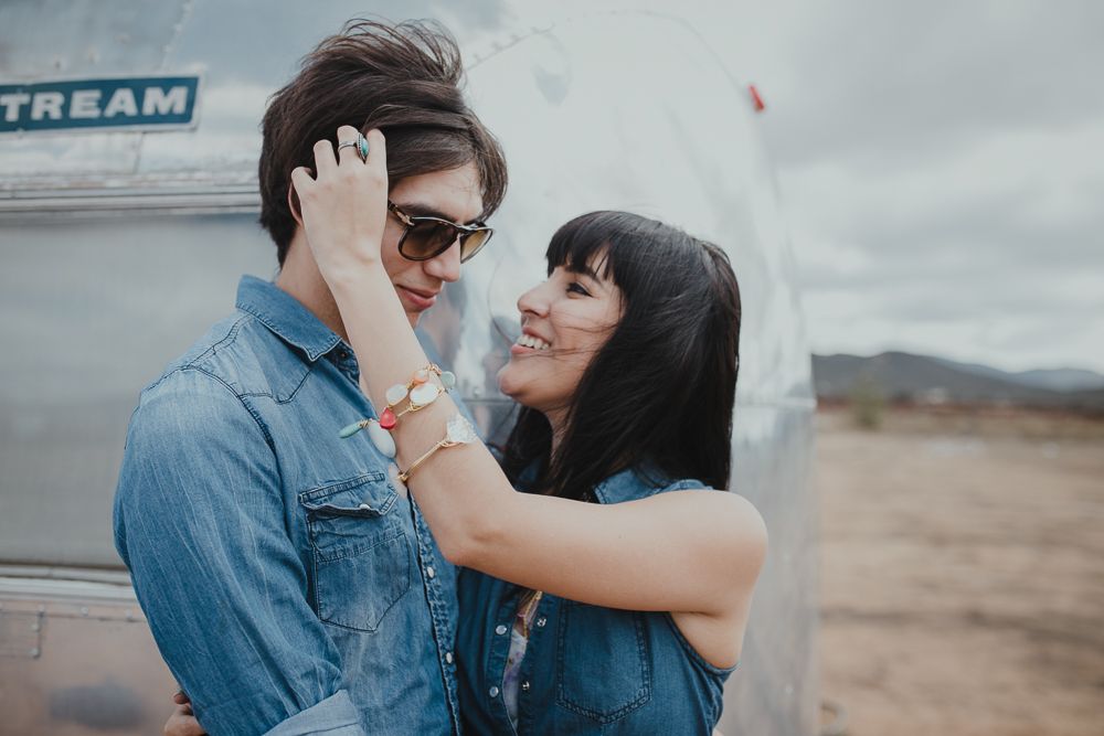 A romantic couple embrace beside an Airstream trailer in a desert setting under cloudy skies.