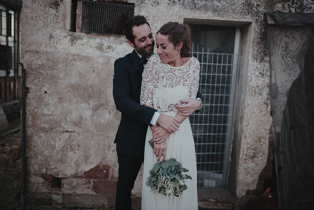 Couple in wedding attire pose against rustic wall in moody wedding photo.