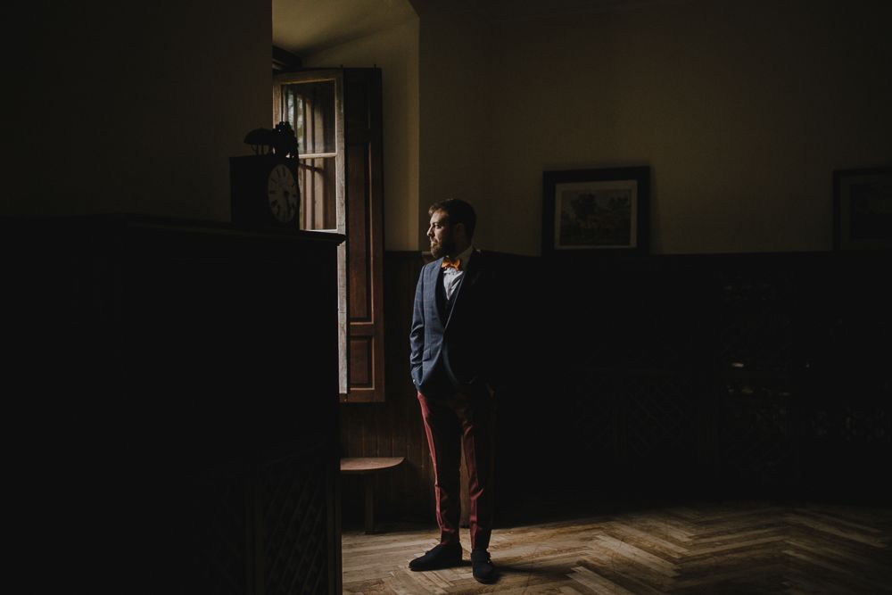 Person in formal attire stands alone in dark room with window light.