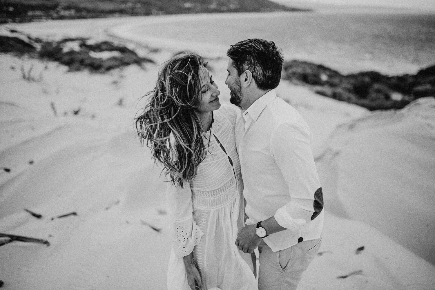Black and white photo of a couple sharing an intimate moment on windswept beach dunes.