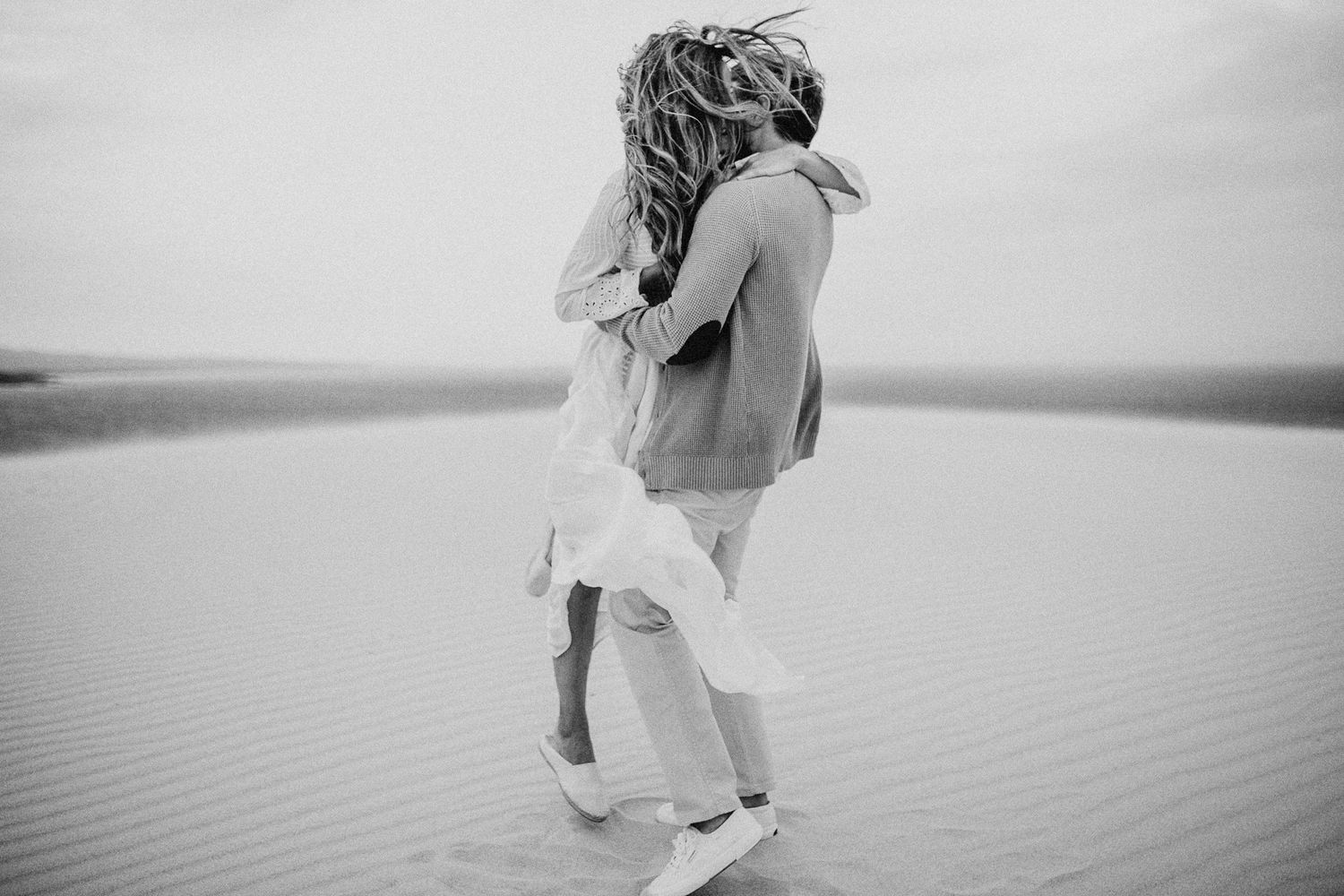 A romantic couple embraces on sandy dunes against an overcast sky in black and white photography.