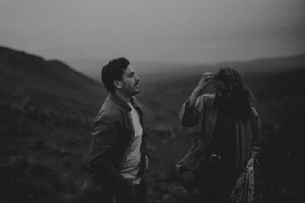 Couple sharing tender moment while perched on rocky outcrop with mountain vista behind them.