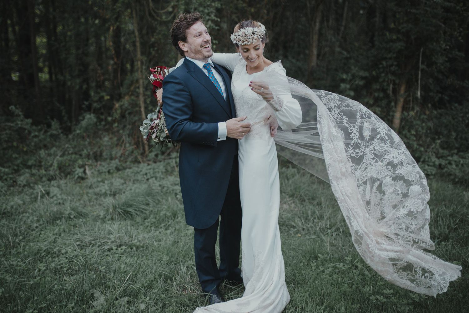 Romantic wedding couple embrace as veil flows in breeze against forest backdrop.