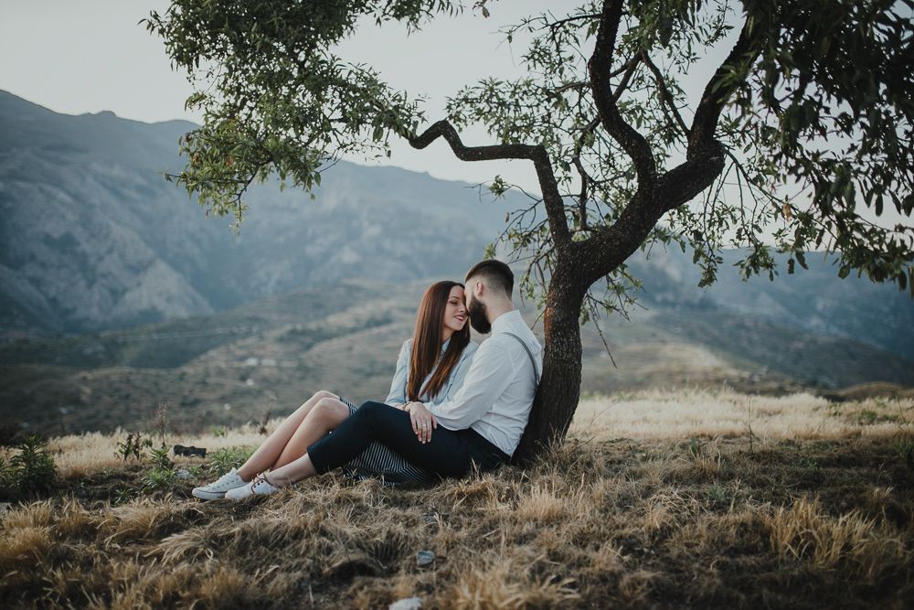 Couple resting under a lone tree on hillside overlooking mountain landscape.