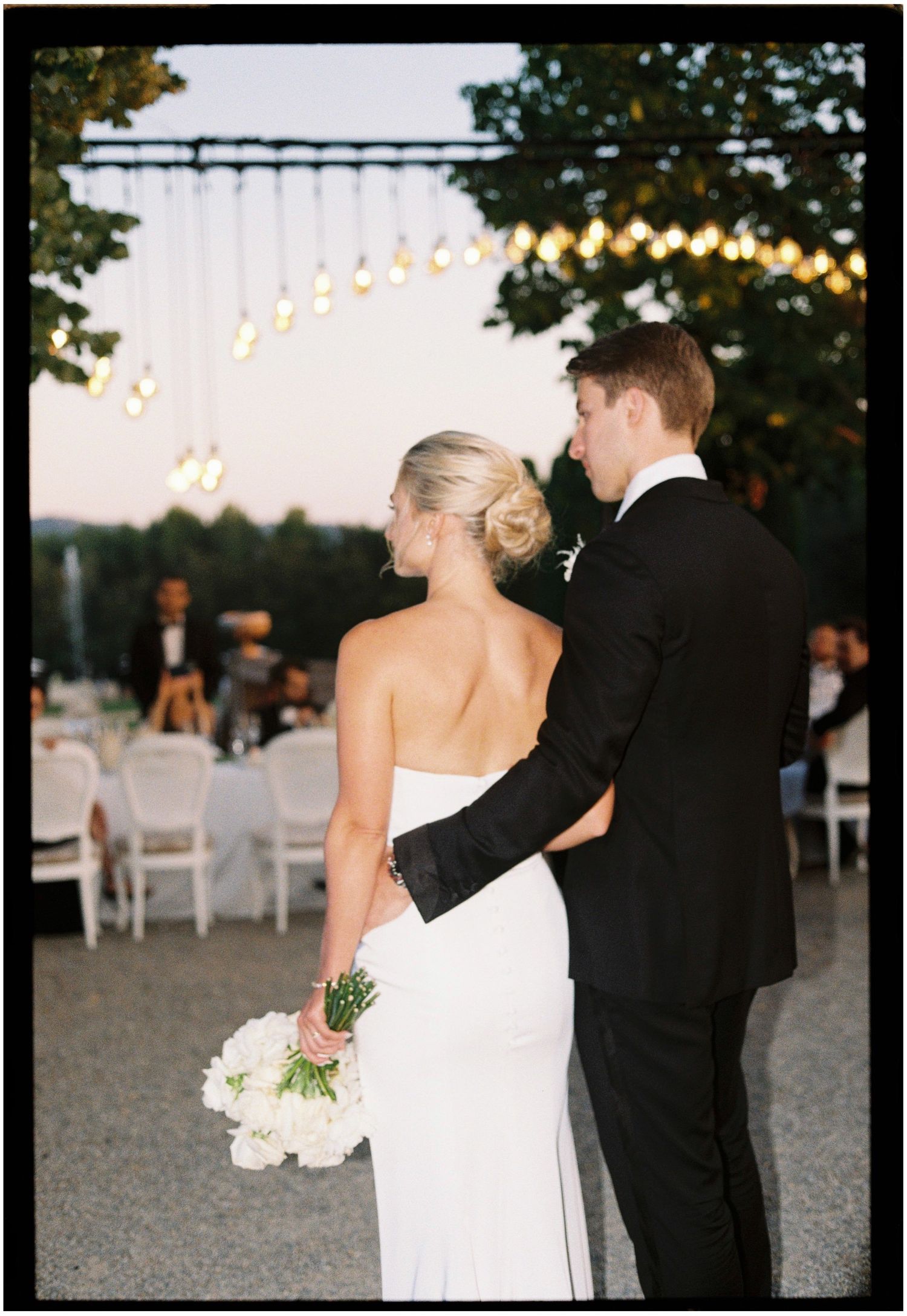 Wedding couple viewing outdoor reception setup with string lights and white chairs.