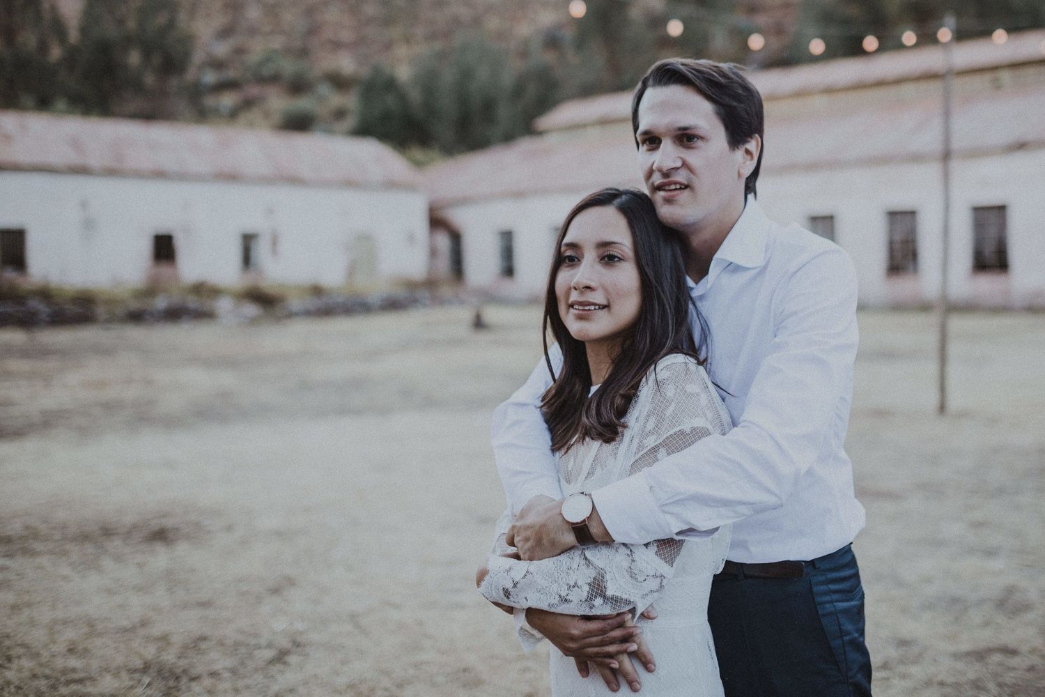 A couple embraces while standing in front of rustic buildings at sunset.