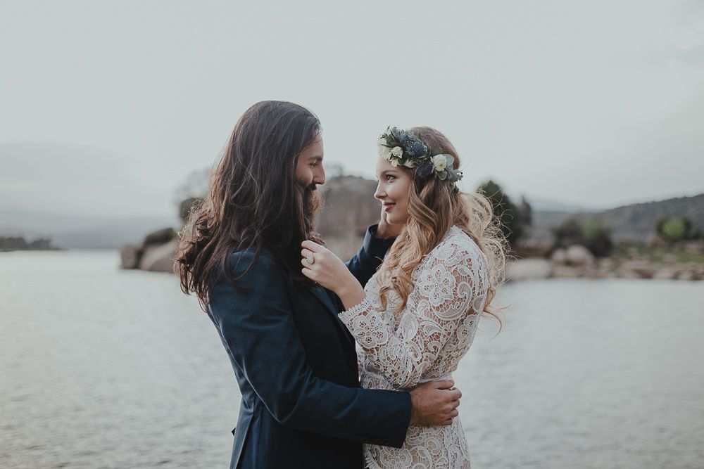 Joyful wedding couple laughs together by mountain lake on cloudy day.