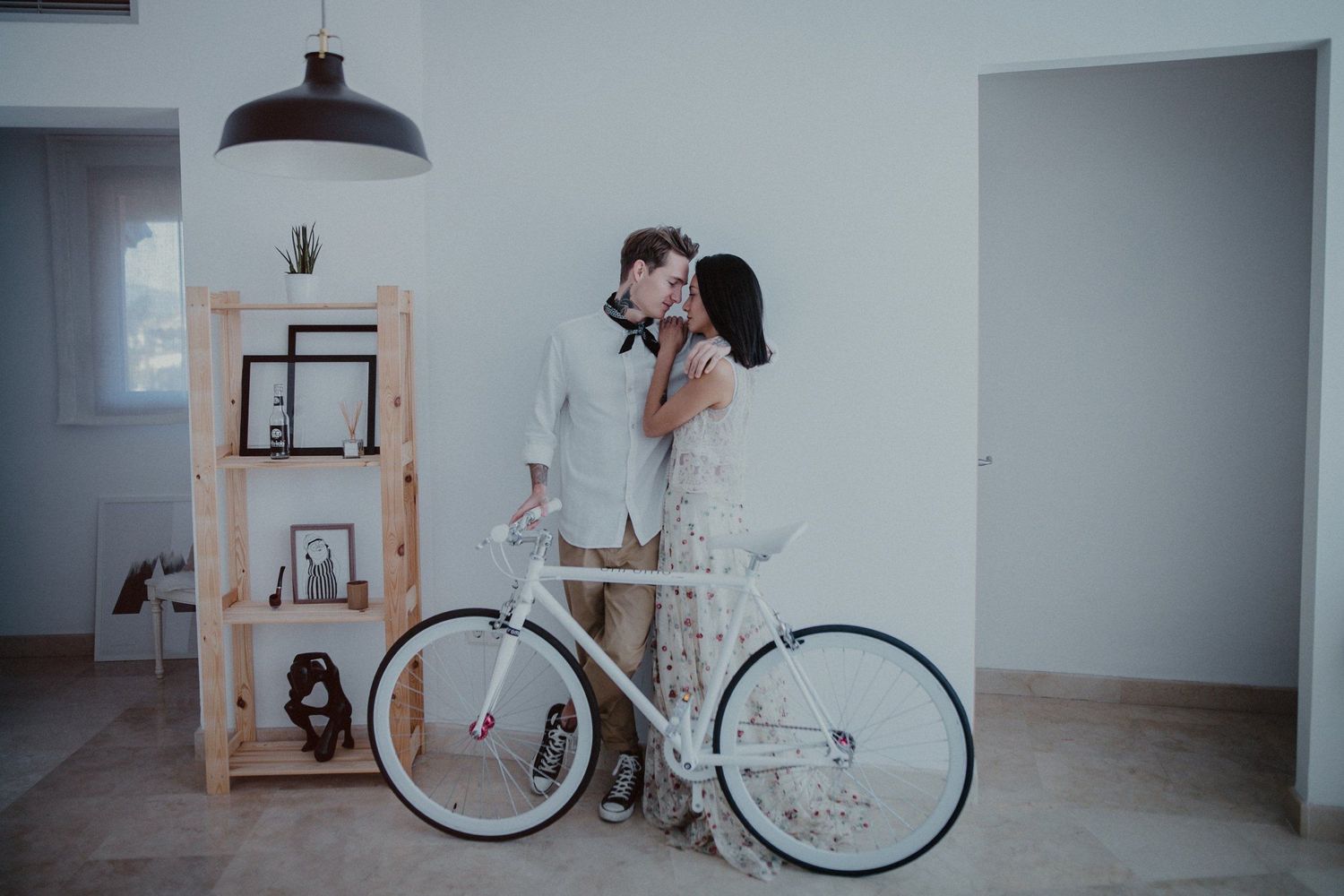 A romantic couple stands together in a minimalist interior with a white bicycle and wooden shelving unit.