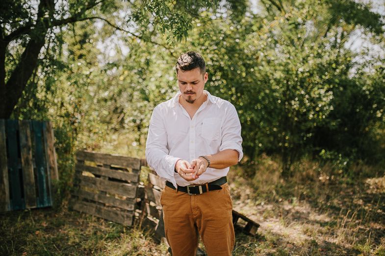 A person in a white shirt and brown pants stands outdoors in front of greenery and wooden crates.