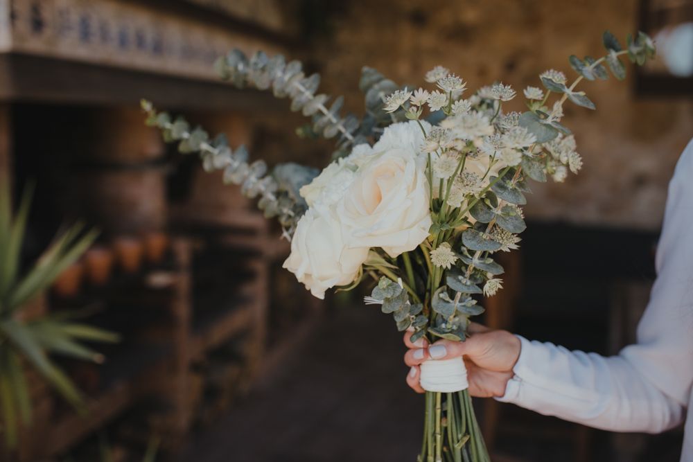 A delicate white rose and eucalyptus bouquet being held by someone in a long-sleeved white garment.