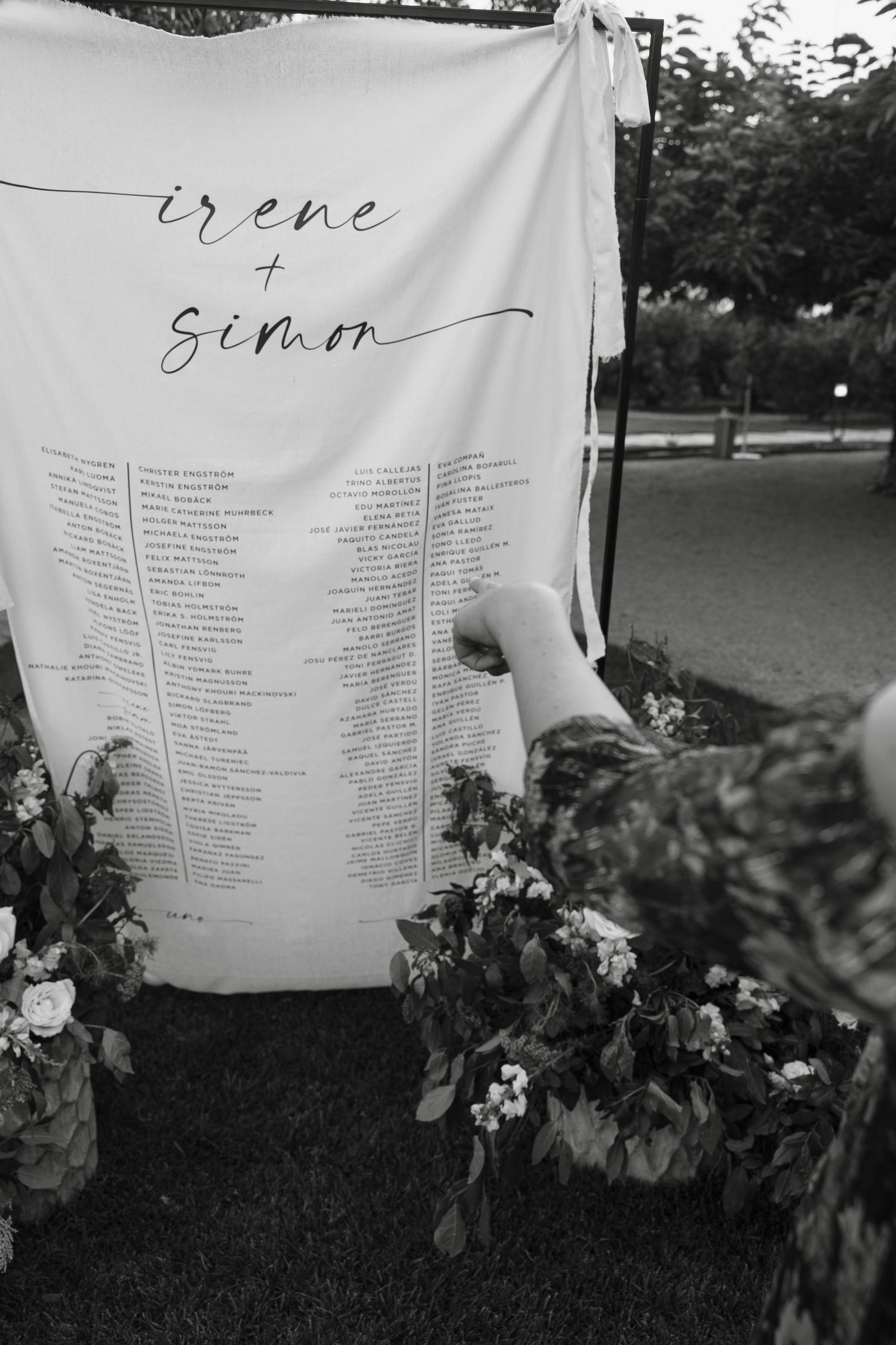 A wedding seating chart banner with calligraphy names and guest list displayed outdoors.