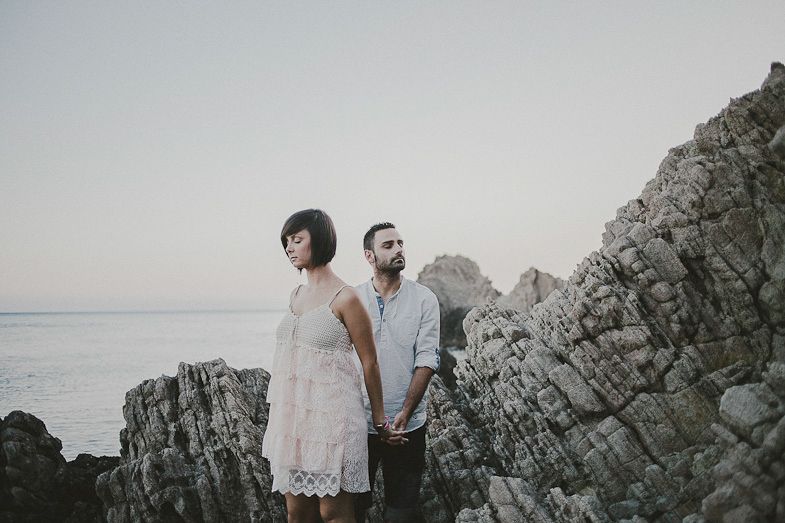 A couple poses dramatically on rocky coastal cliffs at sunset.