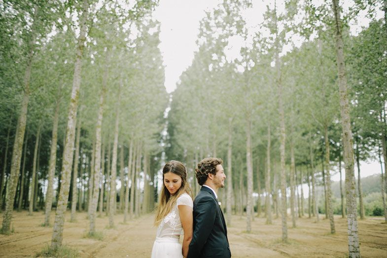 Romantic wedding portrait of couple standing back-to-back among tall forest trees.