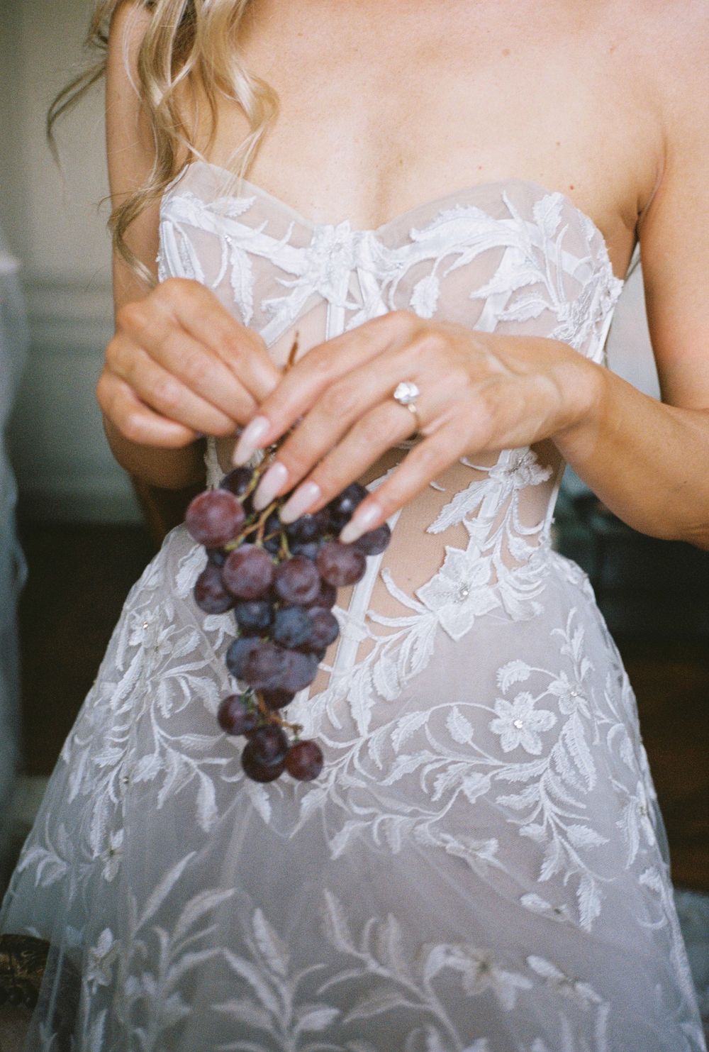 A close-up of lace wedding dress details with purple grapes held against the fabric.