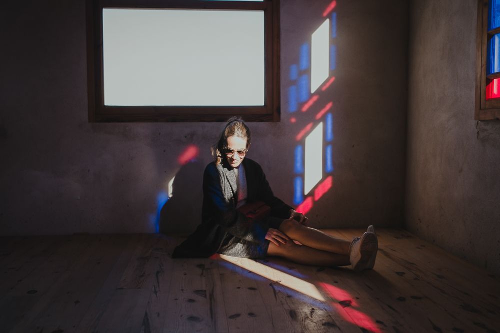 A person in dark clothing sits on a wooden floor with dramatic red and blue light streaming through windows.