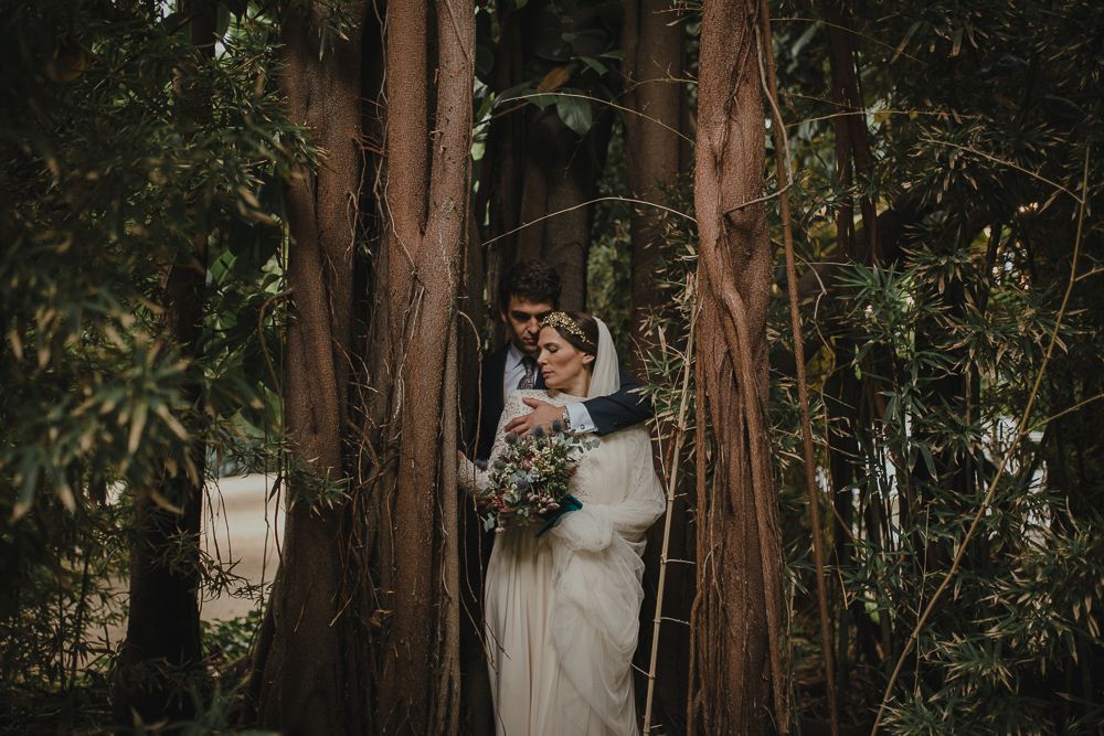 A wedding couple shares an intimate moment among tall trees in a forest setting.