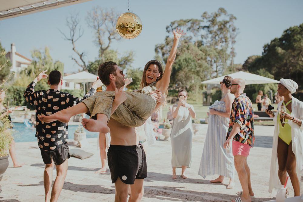 Partygoers dance joyfully at an outdoor pool celebration with a disco ball overhead.
