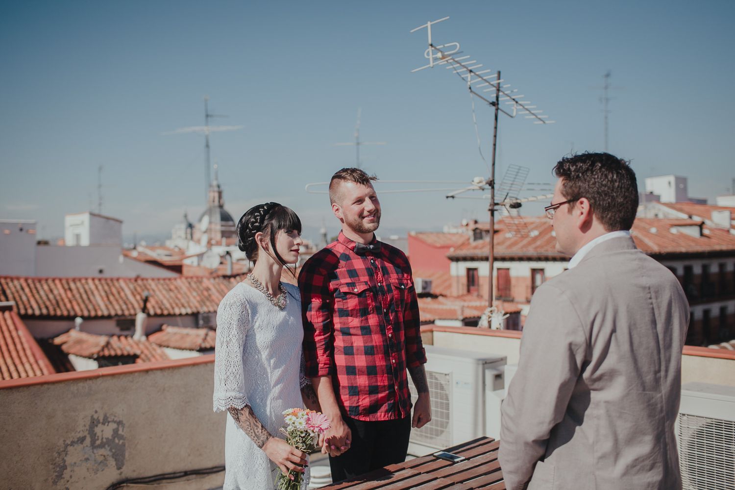 Three people having a conversation on a sunny rooftop with terracotta tiles and antennas visible in the distance.