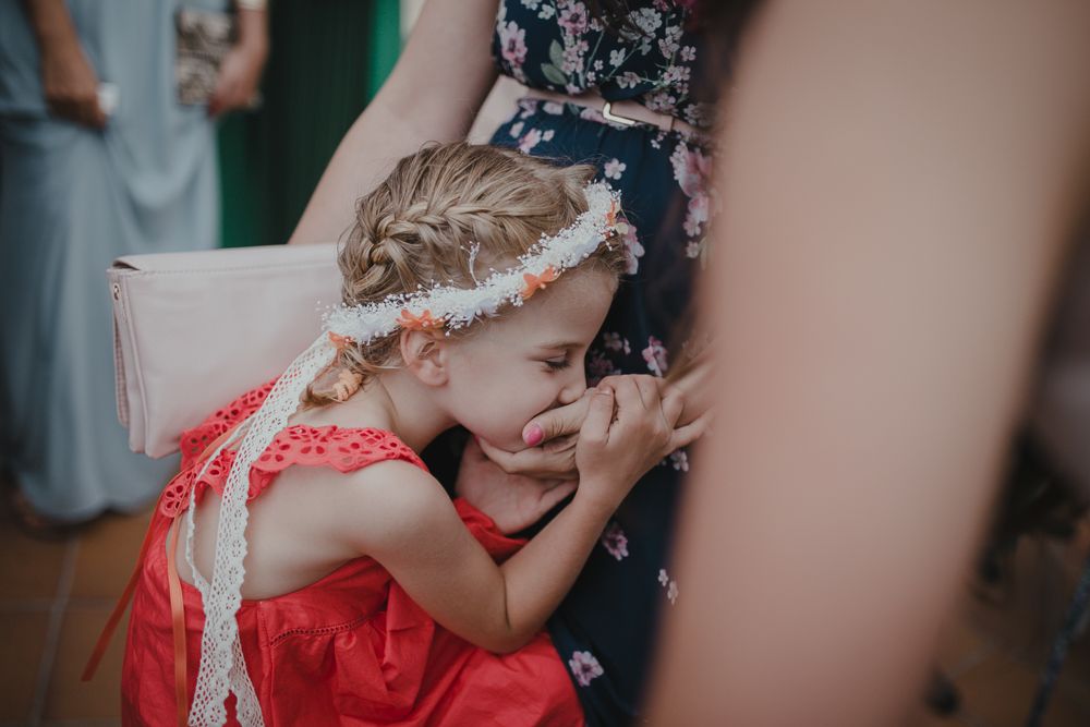 A child in a red dress with a floral headpiece touches something while standing near adults.