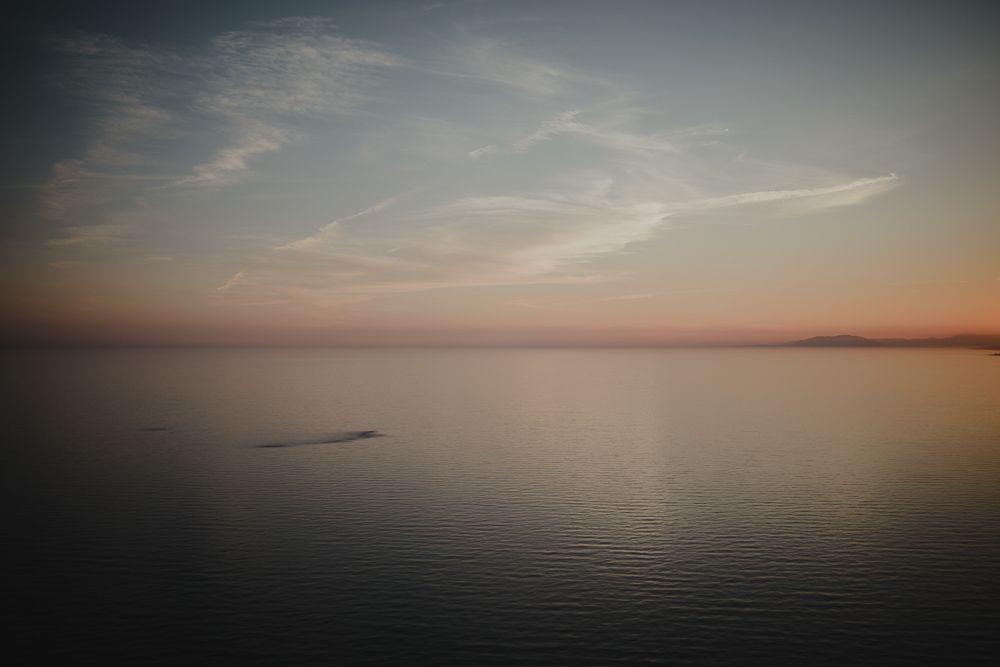 Calm ocean waters reflect a pastel sunset sky with wispy clouds on the horizon.