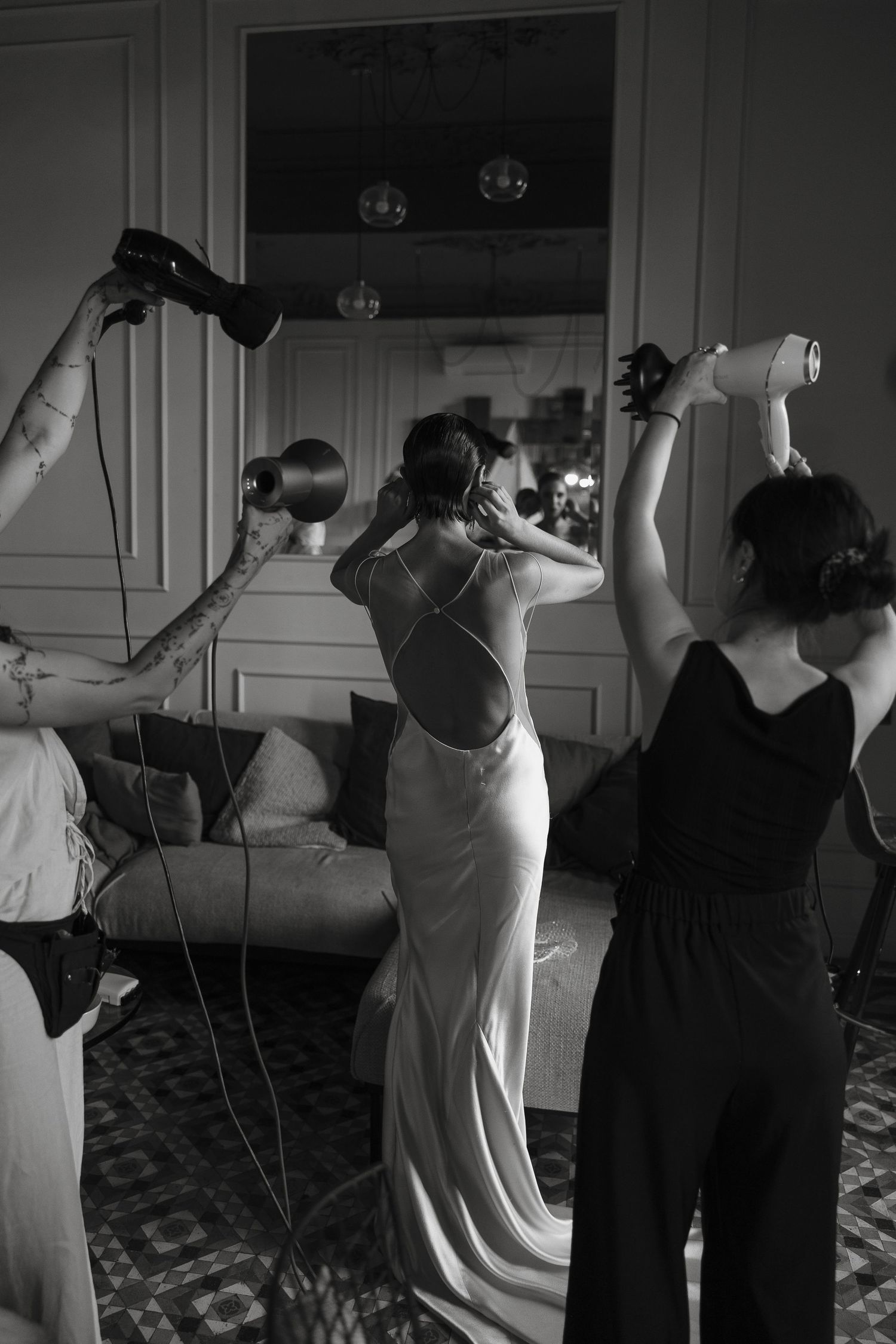 Stylists blow dry brides hair with multiple dryers as she prepares in elegant room before wedding ceremony.