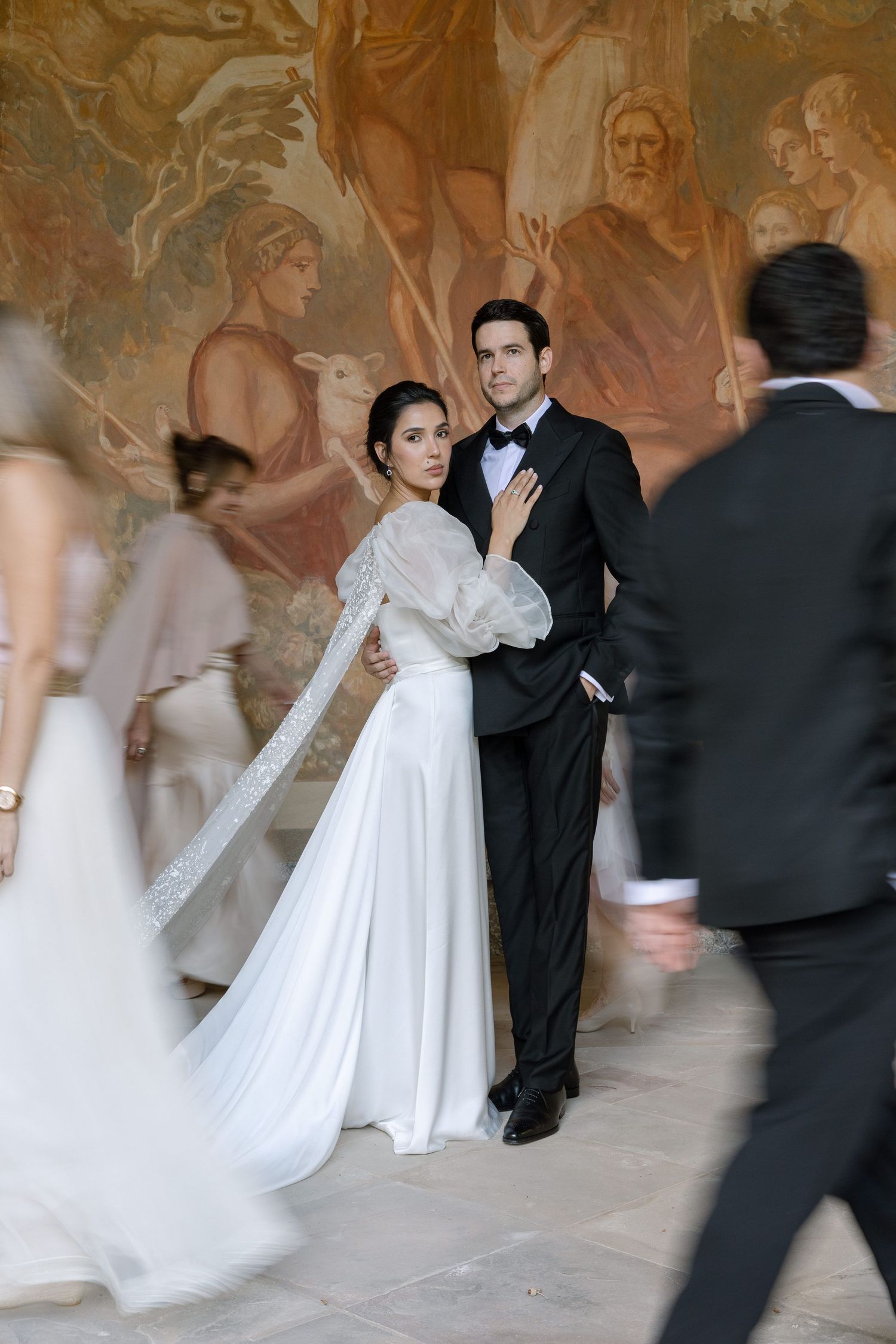 A bride in a flowing white gown dances with guests in formal attire against an ornate wall with decorative murals.