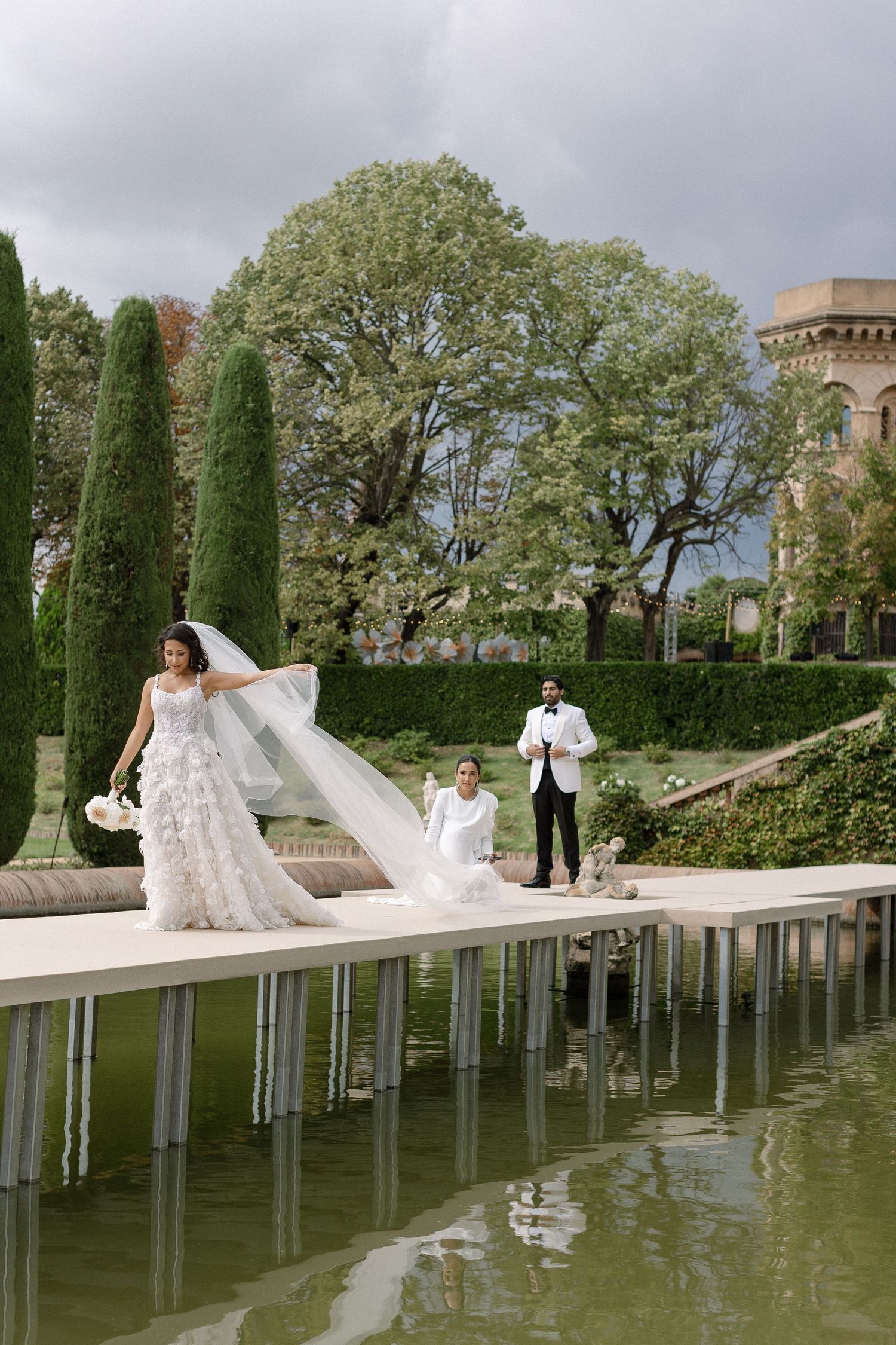 Bride in floral gown with flowing veil walks along reflective pool in elegant garden with cypress trees and historic castle.