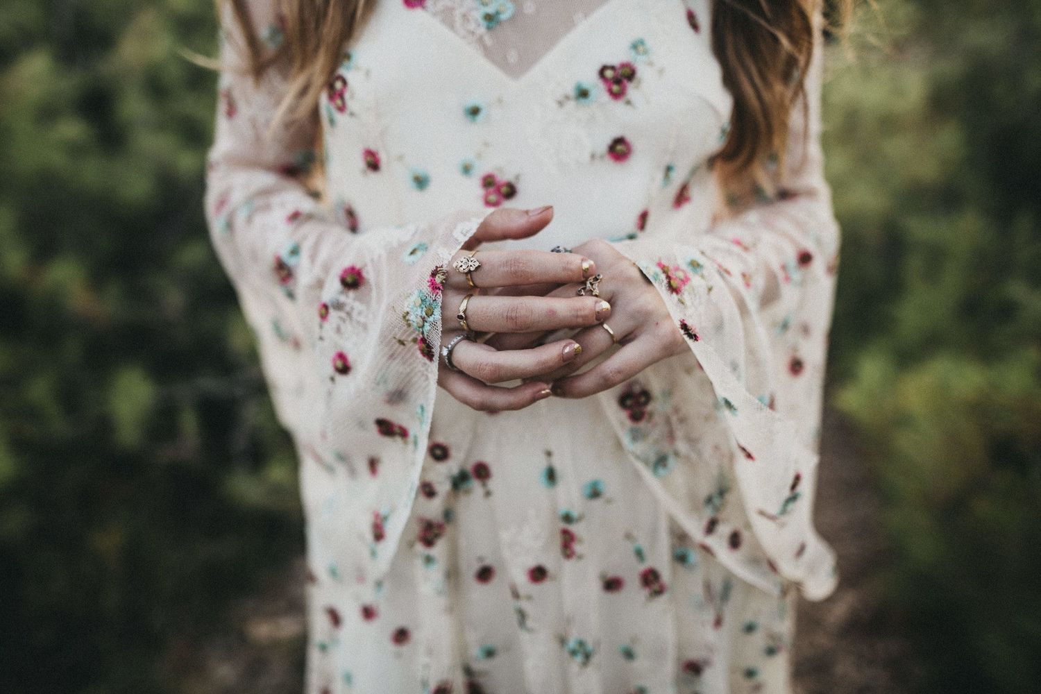 Hands clasped together wearing a flowing white floral dress with bell sleeves against a natural backdrop.