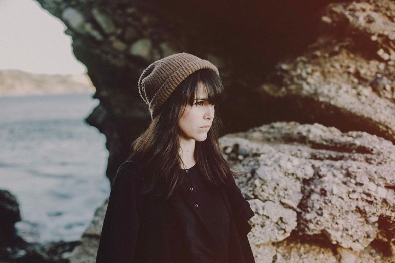 Person in a beanie and dark coat poses near coastal rocks at sunset.