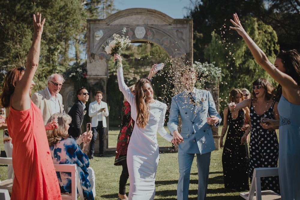 Wedding guests cheer as couple celebrates under stone archway in garden setting.