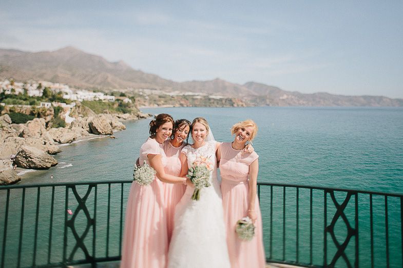 Three people in pink and white dresses pose on a balcony overlooking turquoise Mediterranean waters and rocky coastline.