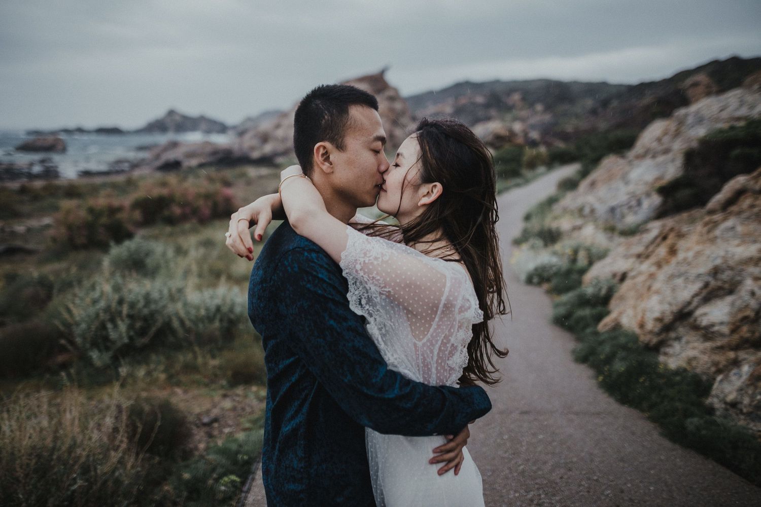 Romantic embrace on coastal path with mountain landscape backdrop.
