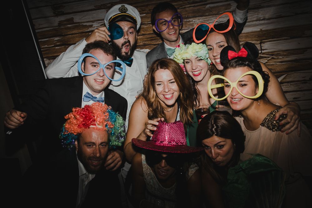 Group of party guests posing with colorful props and flowers in photo booth setting.