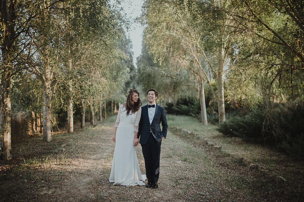 Couple in formal attire standing together on tree-lined path during wedding photoshoot.