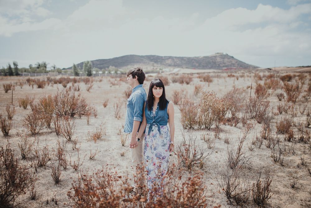 A couple stands together in a desert landscape with scrubby brush and mountains in the background.