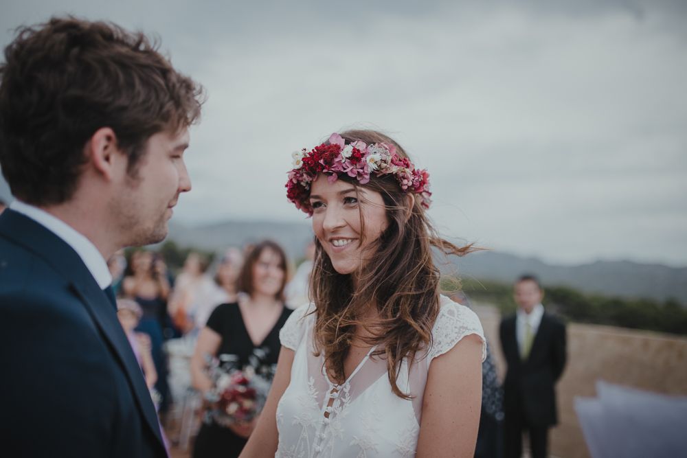 The bride wearing a floral crown and lace dress smiles during an outdoor wedding ceremony.