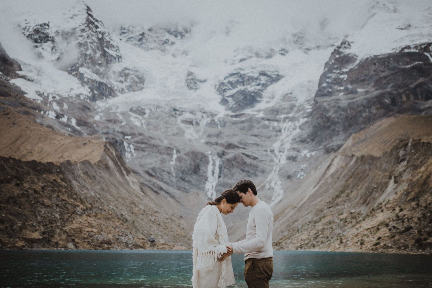 A couple shares an intimate moment before a turquoise glacial lake and snowy mountain peaks.
