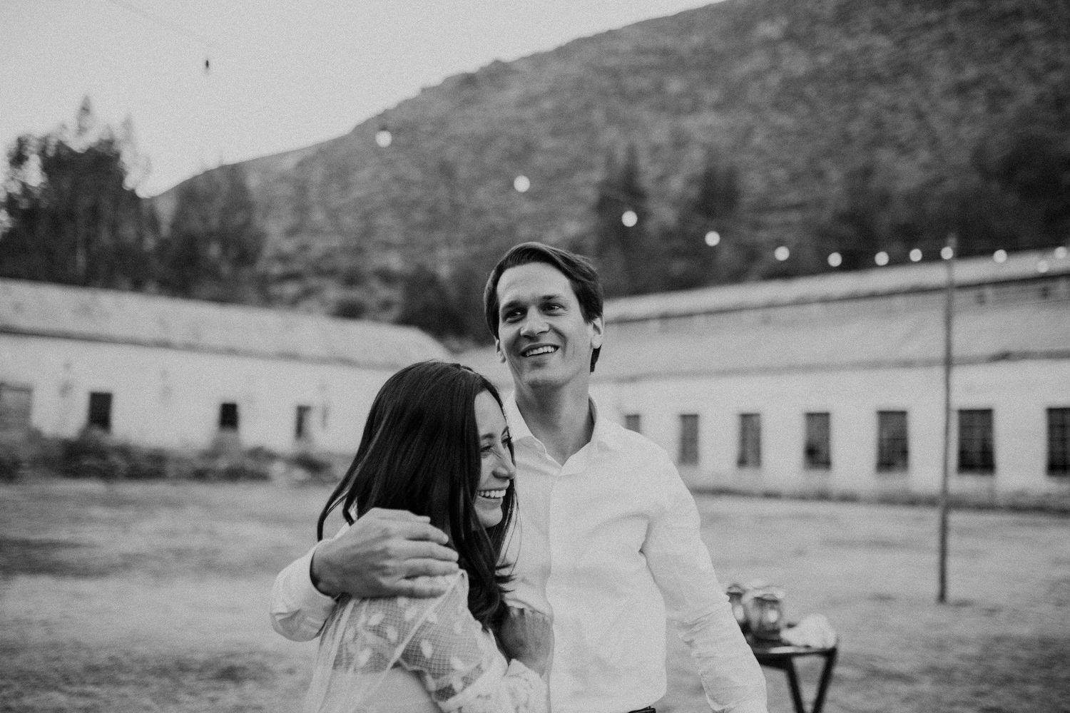 A tender black and white moment of a couple embracing outdoors with white buildings and hills in the background.