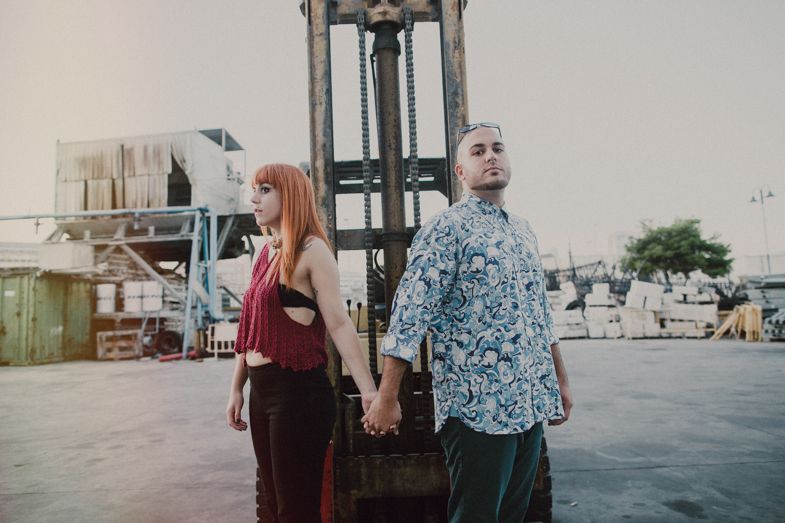 A couple poses in an industrial setting with metal machinery and urban backdrop.