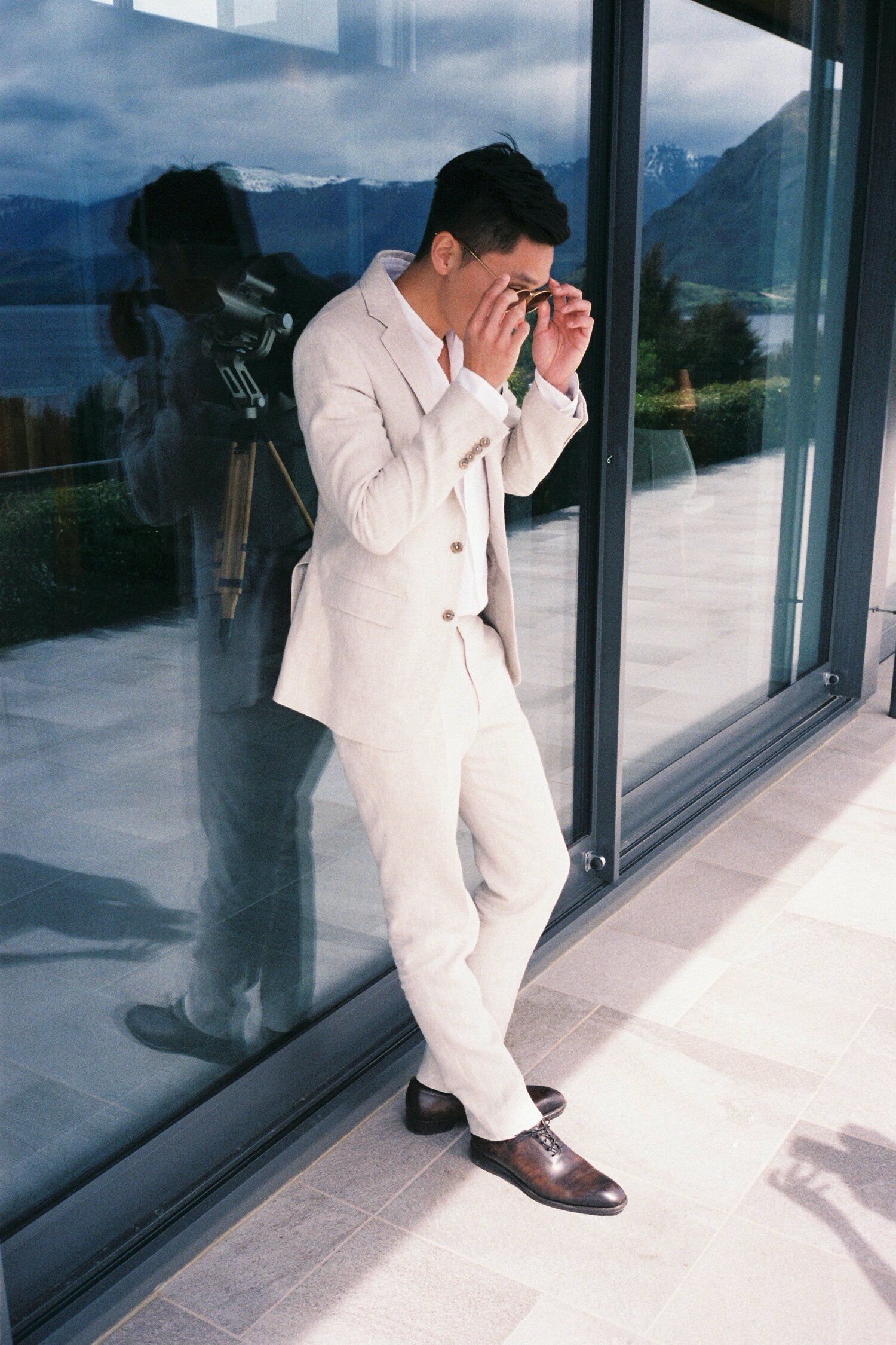 Person in white suit looking through camera near glass window with mountain reflection.